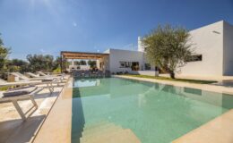 Modern villa with a swimming pool surrounded by lounge chairs and olive trees, set under a clear blue sky. Ideal for luxury vacation rentals or outdoor gatherings.