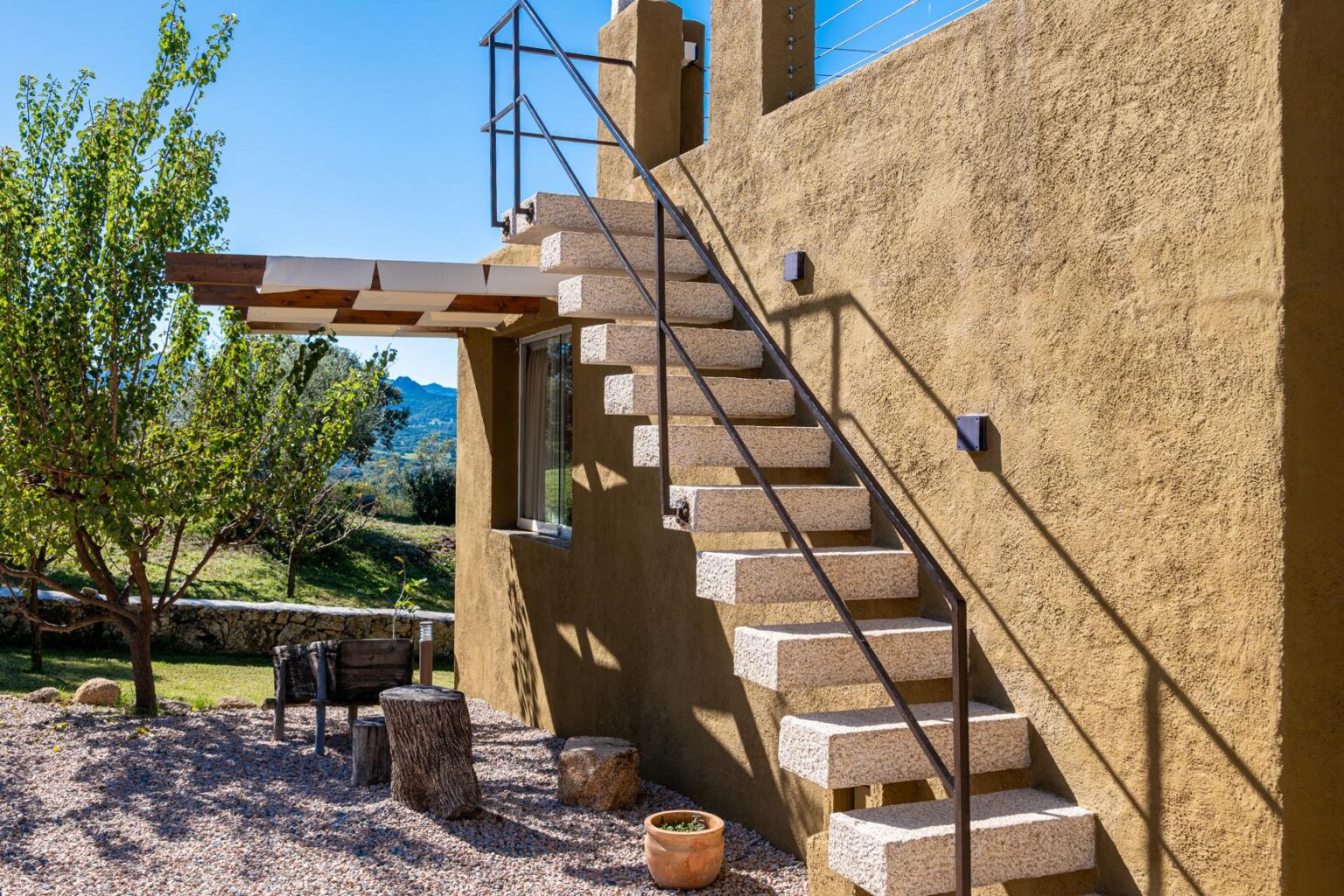 Exterior view of a modern home featuring a stone staircase, surrounded by greenery and outdoor seating, under a clear blue sky. | Only on SopranoVillas