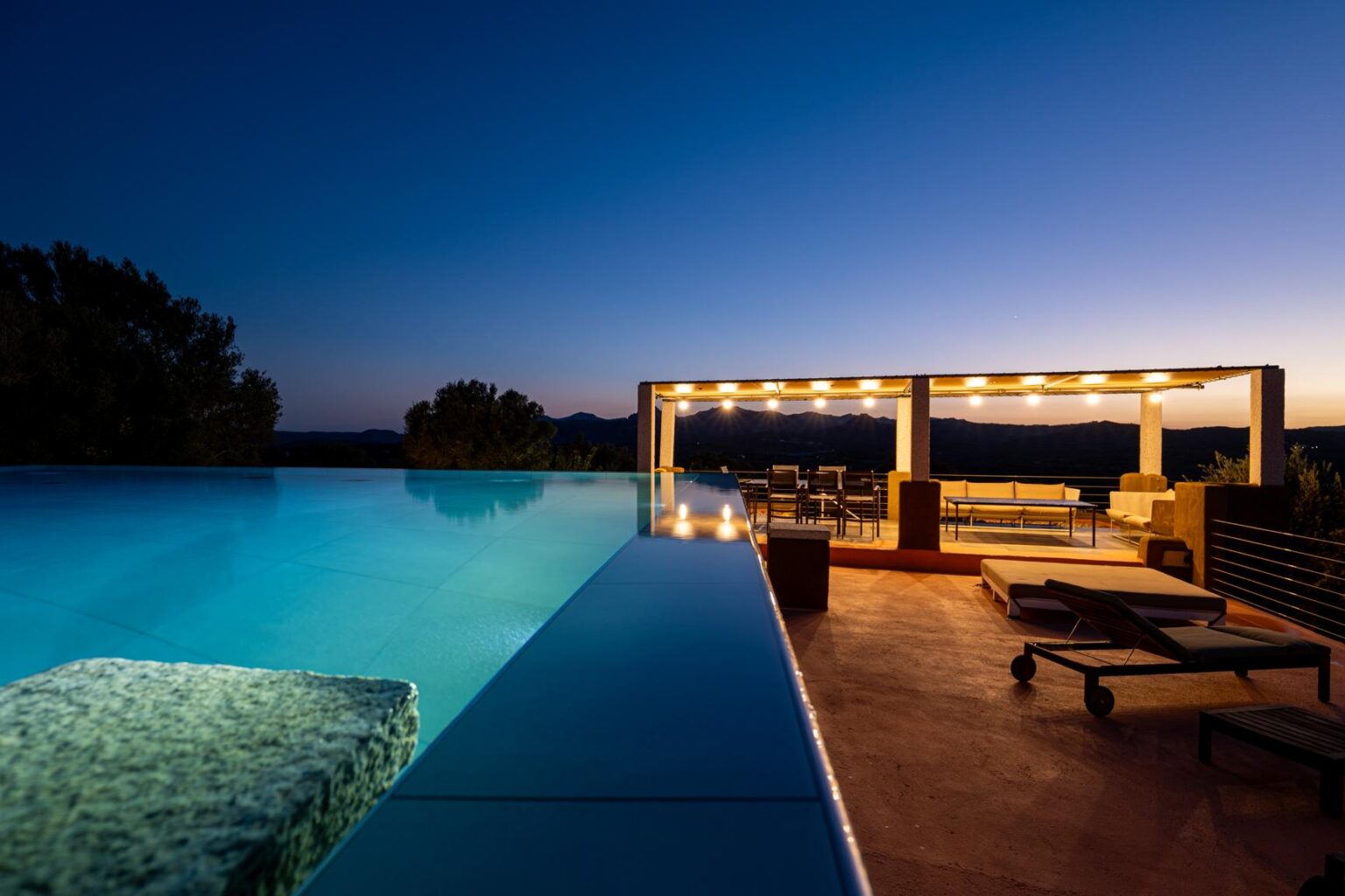 Luxury infinity pool at dusk with a scenic view, surrounded by modern outdoor seating and ambient lighting in a stylish patio setting. | Only on SopranoVillas