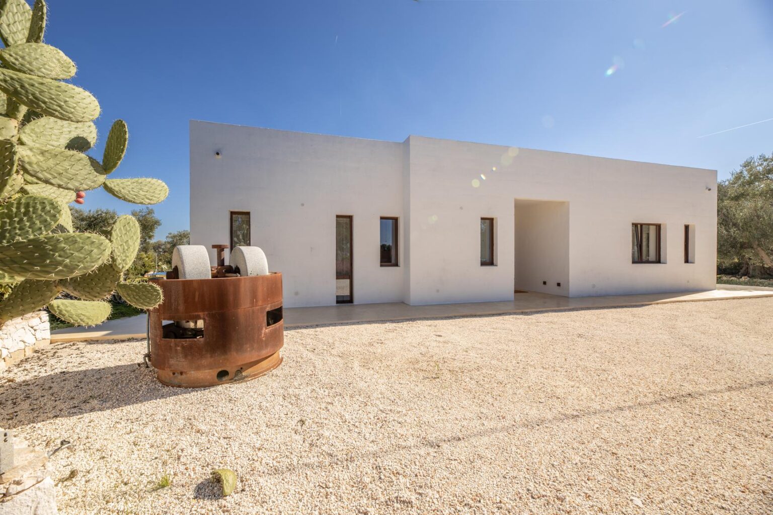 Modern white house with large windows, surrounded by gravel and a cactus plant, featuring a vintage industrial roller in the foreground under a clear blue sky. | Only on SopranoVillas