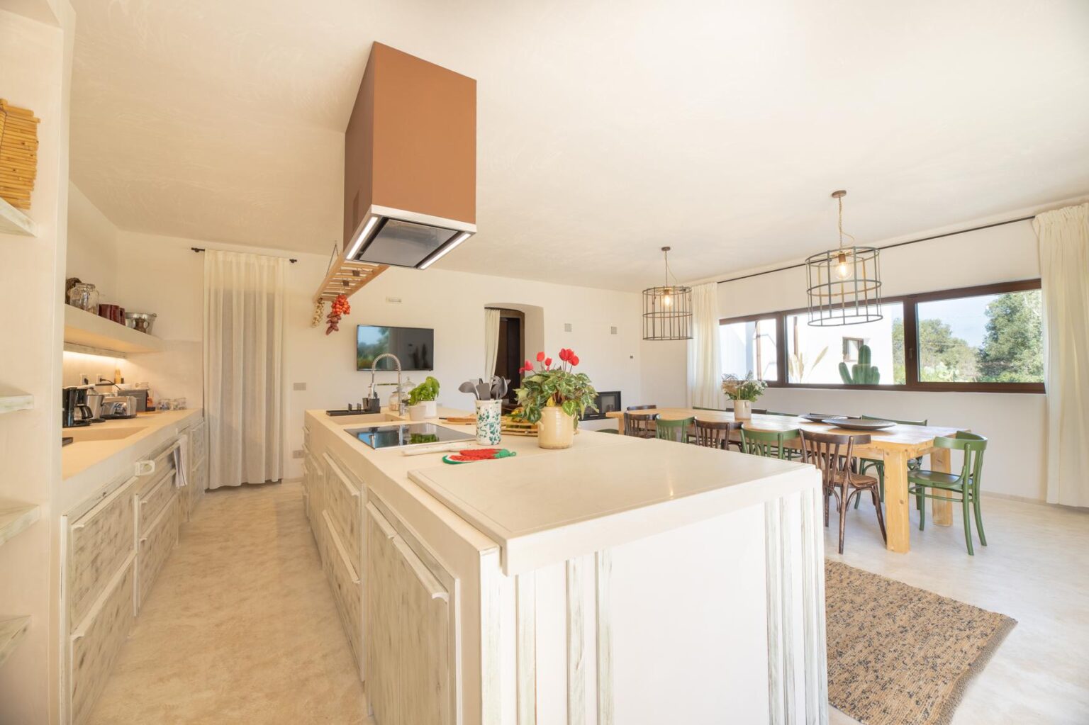 Modern kitchen interior with white cabinetry, a spacious island, and a dining area featuring wooden tables and colorful chairs. Natural light floods the space through large windows, creating a bright and inviting atmosphere. | Only on SopranoVillas