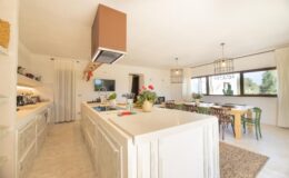 Modern kitchen interior with white cabinetry, a spacious island, and a dining area featuring wooden tables and colorful chairs. Natural light floods the space through large windows, creating a bright and inviting atmosphere.