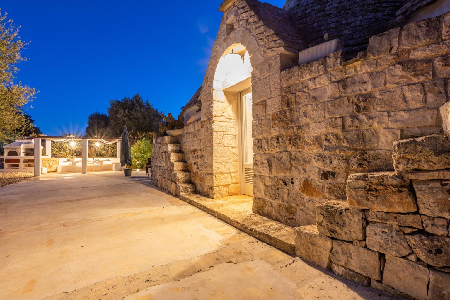 Stone cottage entrance illuminated at dusk, showcasing rustic architecture and a welcoming pathway. Lush greenery and warm outdoor lighting enhance the inviting atmosphere. | Only on SopranoVillas