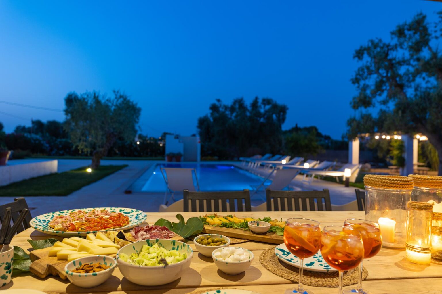 Outdoor dining setup at dusk featuring a variety of gourmet dishes, including fresh salads, cheeses, and fruits, with a beautifully lit swimming pool and landscaped garden in the background. Perfect for evening gatherings and summer entertaining. | Only on SopranoVillas