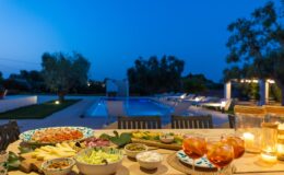 Outdoor dining setup at dusk featuring a variety of gourmet dishes, including fresh salads, cheeses, and fruits, with a beautifully lit swimming pool and landscaped garden in the background. Perfect for evening gatherings and summer entertaining.