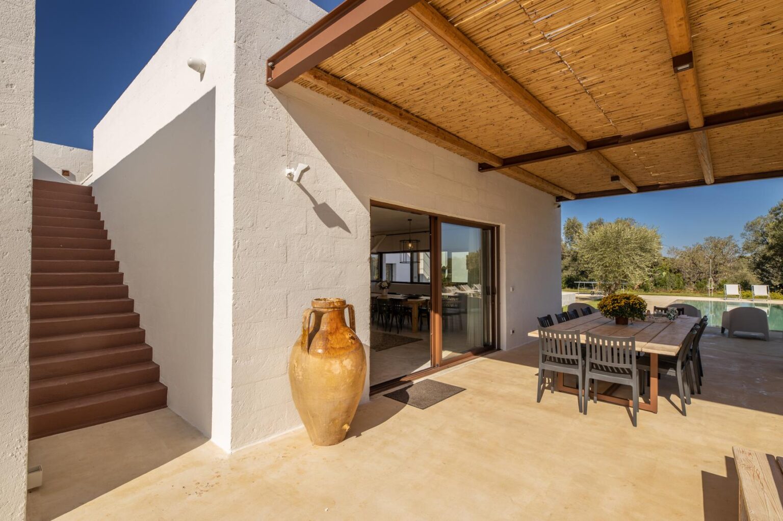 Modern outdoor living space featuring a staircase, large glass doors, and a rustic ceramic vase, surrounded by a sunny landscape and shaded dining area under a thatched roof. | Only on SopranoVillas