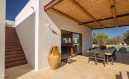 Modern outdoor living space featuring a staircase, large glass doors, and a rustic ceramic vase, surrounded by a sunny landscape and shaded dining area under a thatched roof.