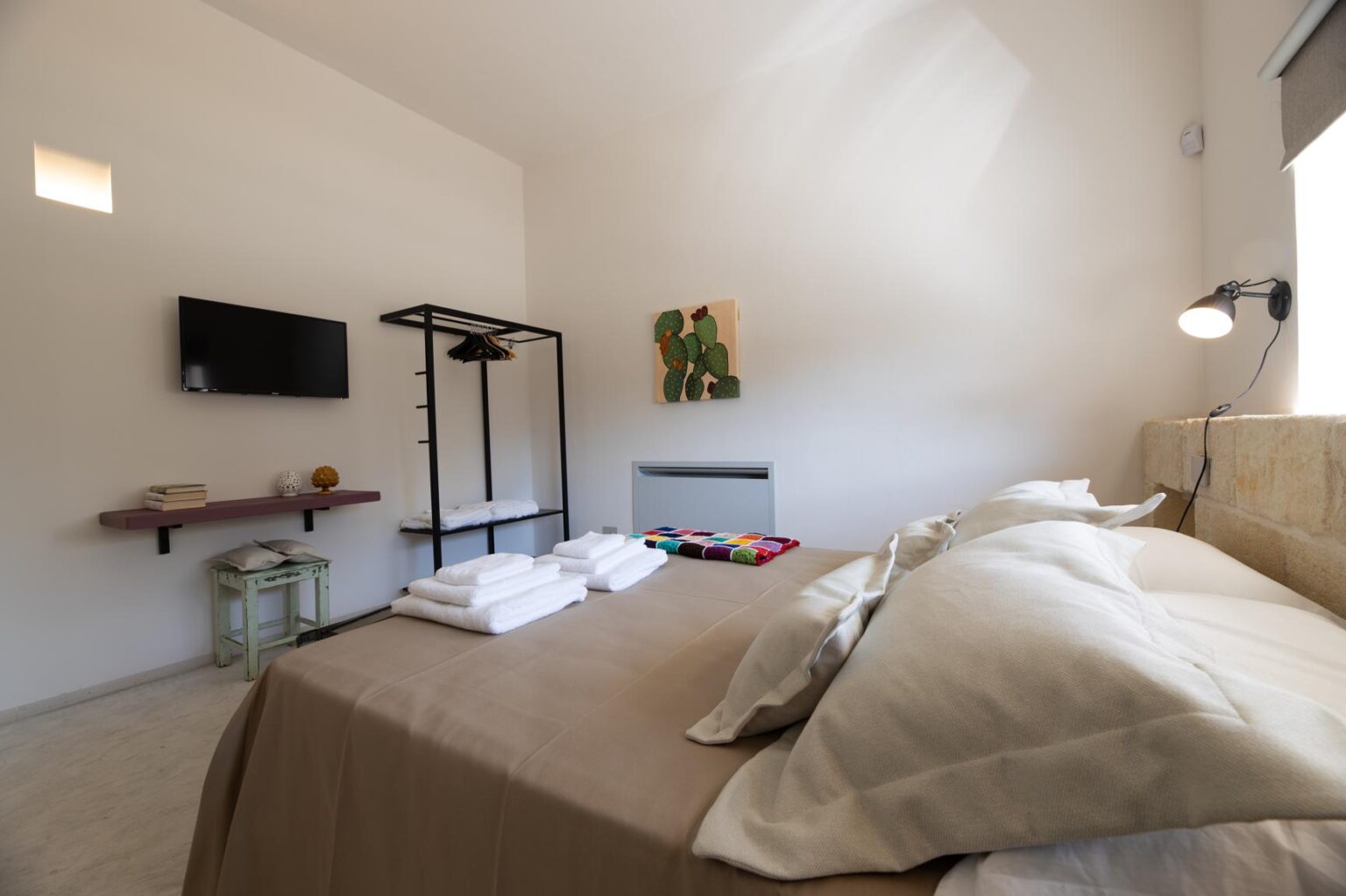 Cozy bedroom interior featuring a neatly made bed with beige linens and fluffy pillows, a wall-mounted TV, a clothing rack with hangers, and a decorative wall art piece. Soft lighting creates a warm atmosphere, complemented by a small bedside table with books and a decorative object. | Only on SopranoVillas