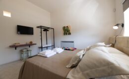 Cozy bedroom interior featuring a neatly made bed with beige linens and fluffy pillows, a wall-mounted TV, a clothing rack with hangers, and a decorative wall art piece. Soft lighting creates a warm atmosphere, complemented by a small bedside table with books and a decorative object.
