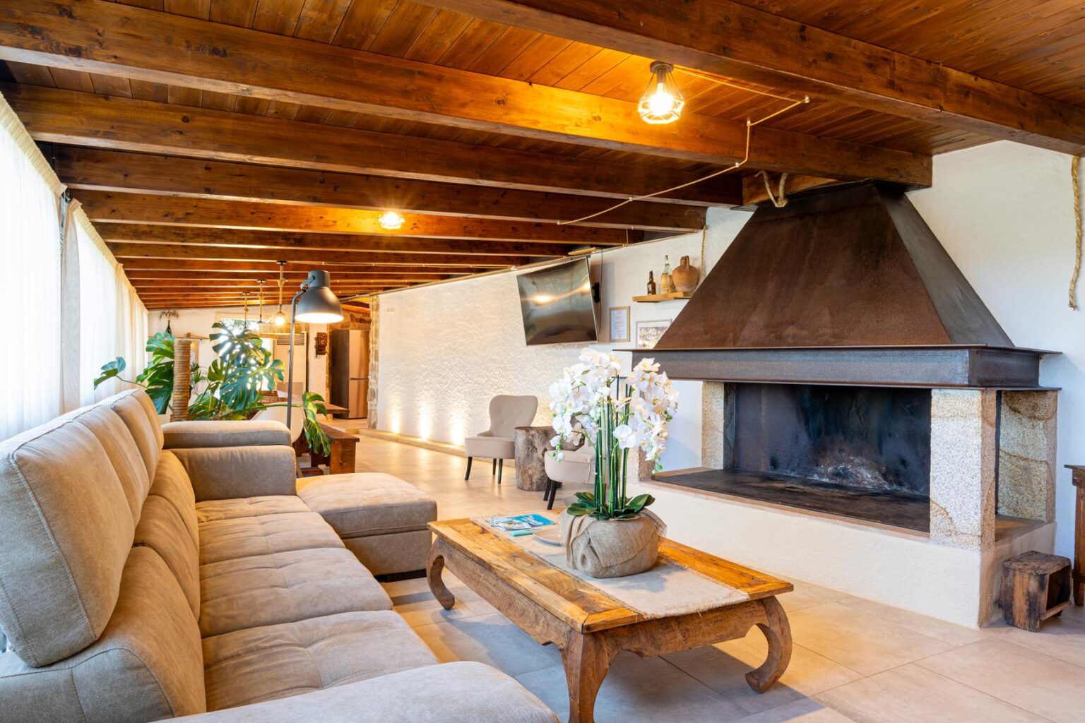 Cozy living room featuring a beige sectional sofa, wooden beams on the ceiling, a stone fireplace, and a wooden coffee table with a potted plant, creating a warm and inviting atmosphere. | Only on SopranoVillas