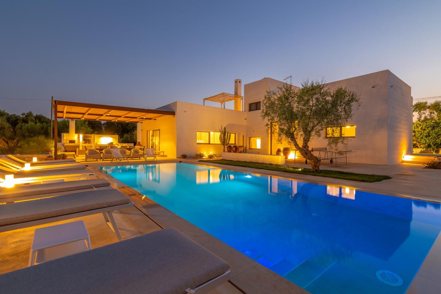 Modern luxury villa with a swimming pool, illuminated at dusk, surrounded by lush greenery and outdoor seating areas. Ideal for relaxation and entertainment. | Only on SopranoVillas