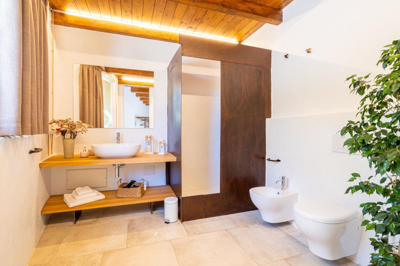 Modern bathroom featuring a wooden ceiling, minimalist decor, a large mirror, a wooden sink vanity with storage, and a stylish toilet, complemented by a potted plant for a touch of greenery. | Only on SopranoVillas