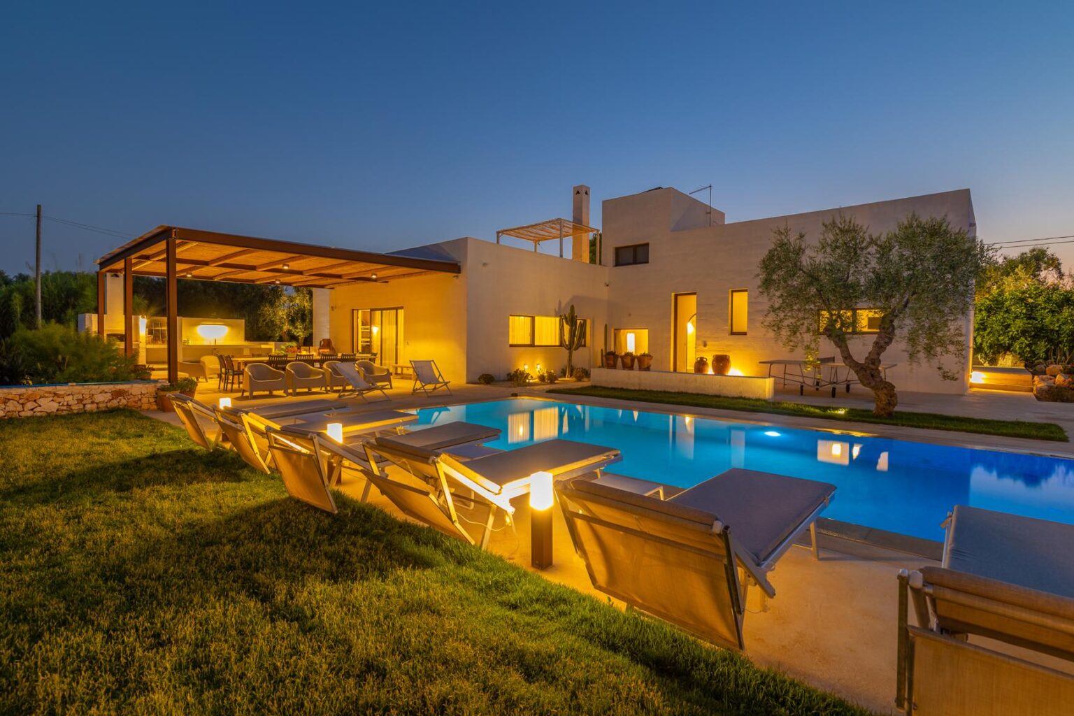 Modern villa with a swimming pool and lounge chairs, illuminated at dusk, surrounded by lush greenery and outdoor seating areas. Ideal for luxury vacation rentals. | Only on SopranoVillas