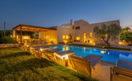 Modern villa with a swimming pool and lounge chairs, illuminated at dusk, surrounded by lush greenery and outdoor seating areas. Ideal for luxury vacation rentals.