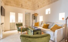 Cozy living room featuring a white sofa with colorful cushions, a green armchair, a wooden coffee table, and natural stone walls, creating a warm and inviting atmosphere.