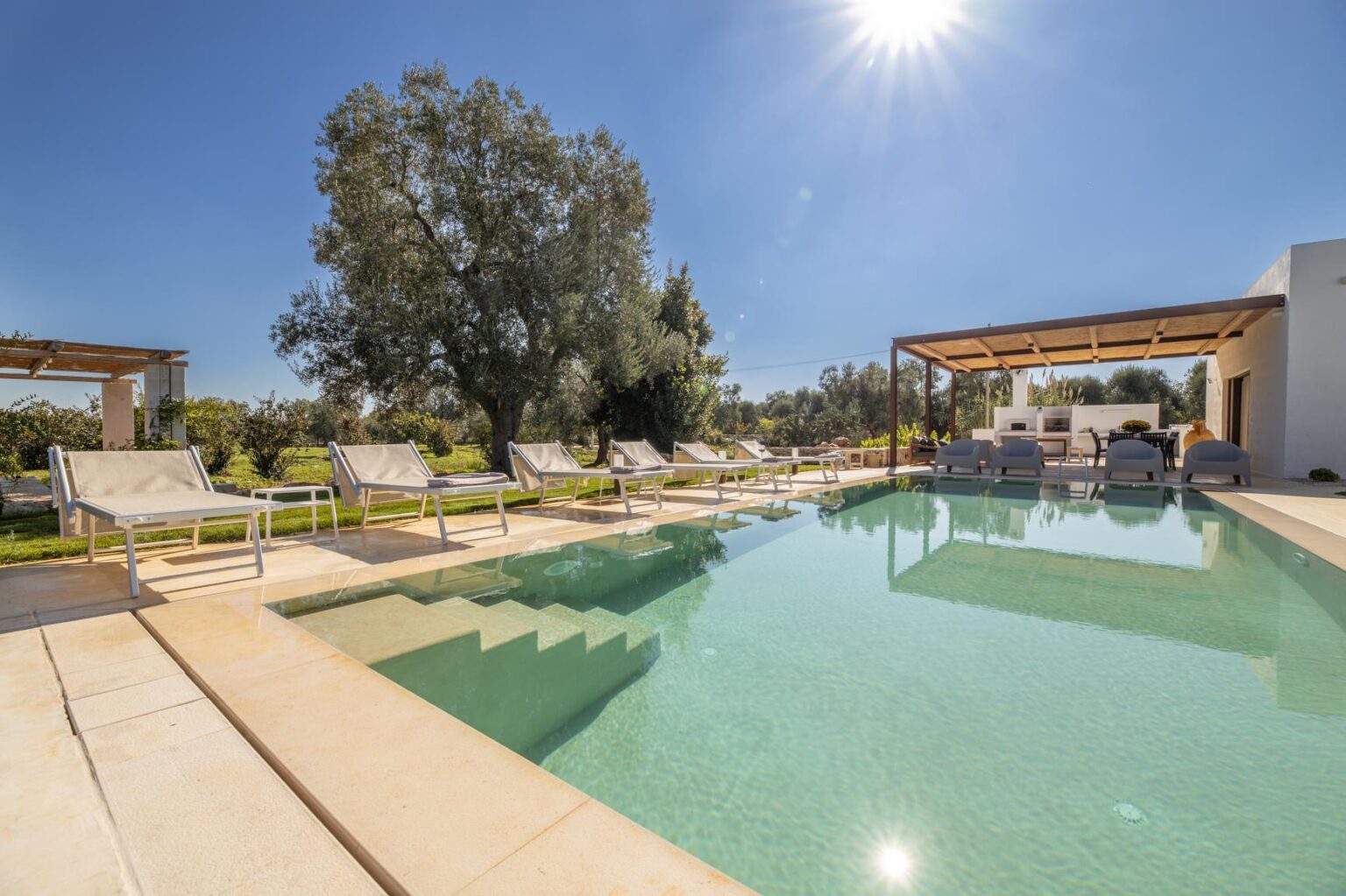 Luxurious outdoor pool area with sun loungers, surrounded by lush greenery and a shaded seating area, under a clear blue sky with bright sunlight. Perfect for relaxation and leisure in a serene environment. | Only on SopranoVillas