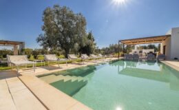 Luxurious outdoor pool area with sun loungers, surrounded by lush greenery and a shaded seating area, under a clear blue sky with bright sunlight. Perfect for relaxation and leisure in a serene environment.