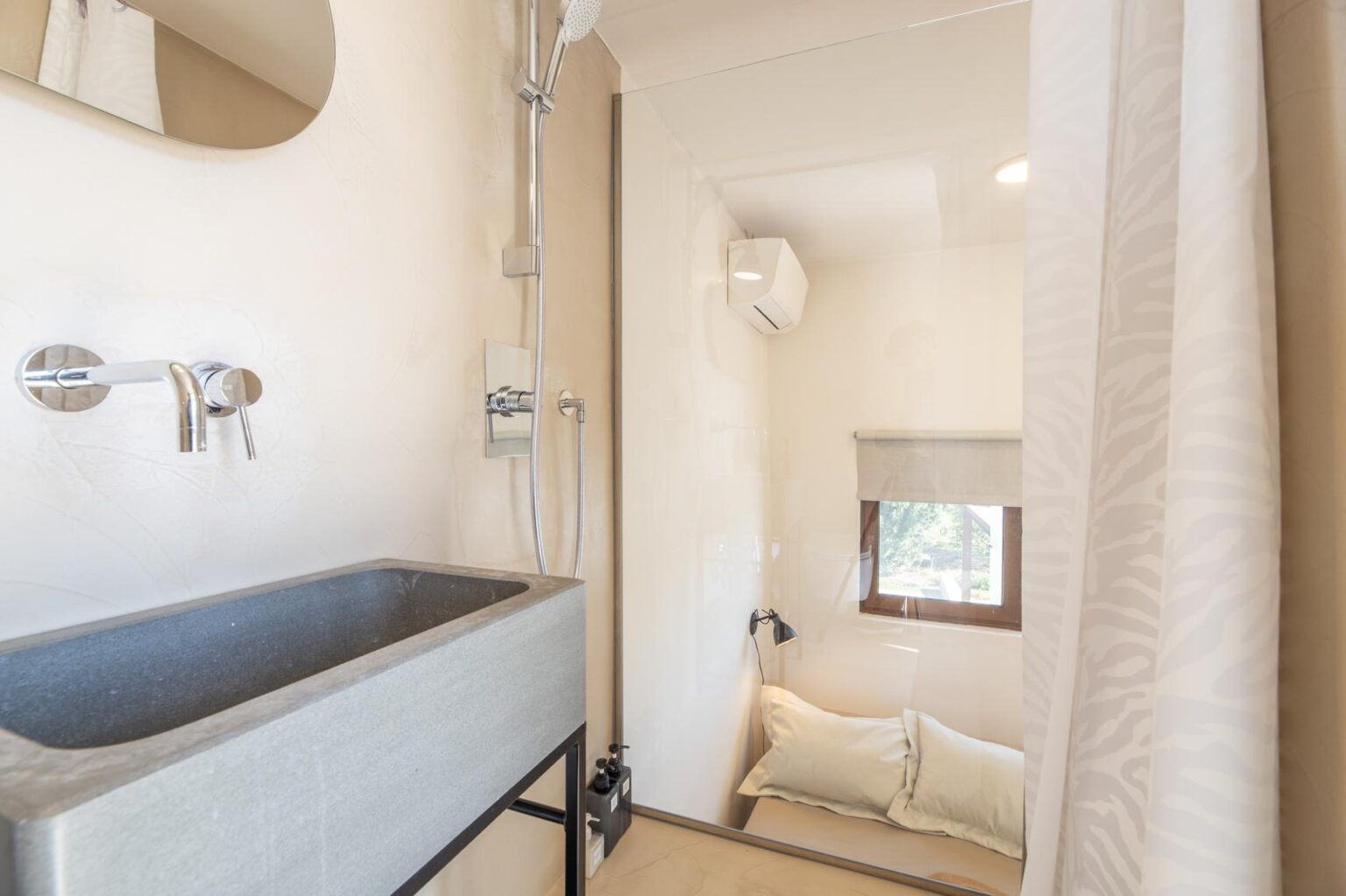 Modern bathroom featuring a sleek gray stone sink, chrome faucets, and a glass shower enclosure. The space includes a cozy nook with pillows, a window for natural light, and an air conditioning unit, creating a relaxing atmosphere. | Only on SopranoVillas