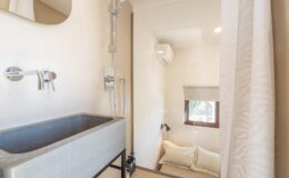 Modern bathroom featuring a sleek gray stone sink, chrome faucets, and a glass shower enclosure. The space includes a cozy nook with pillows, a window for natural light, and an air conditioning unit, creating a relaxing atmosphere.