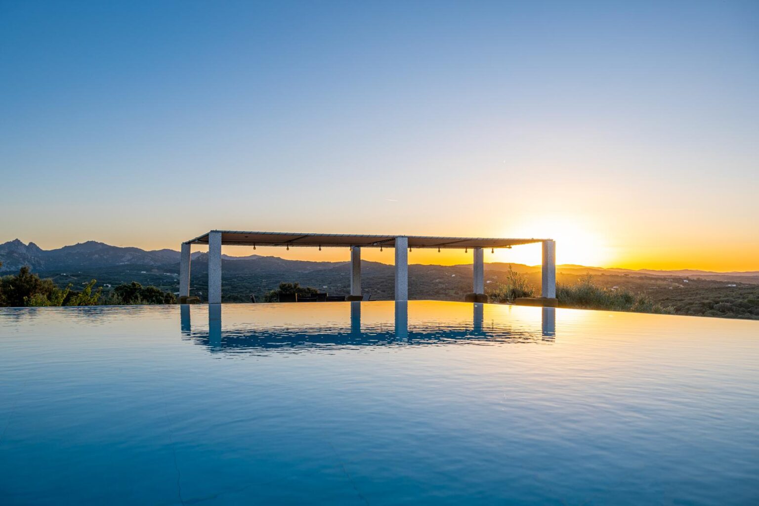 Modern infinity pool reflecting the sunset with a pergola in the background, surrounded by scenic mountains and lush greenery. Ideal for luxury vacation destinations. | Only on SopranoVillas