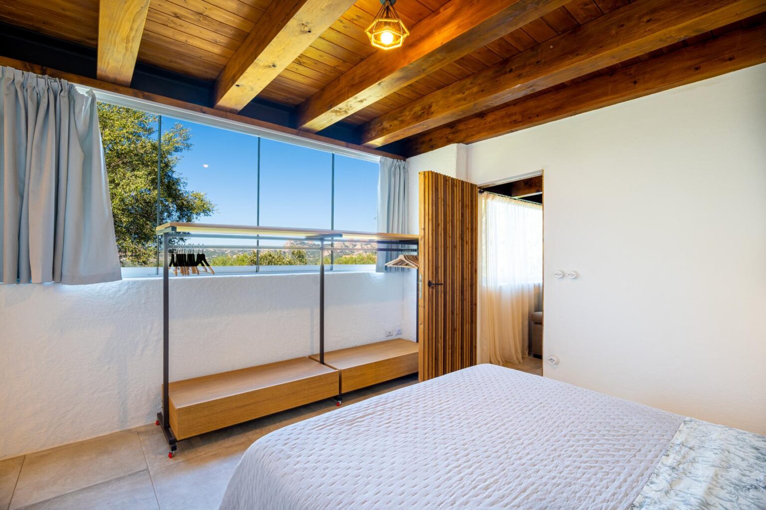 Modern bedroom interior featuring wooden beams, large windows with scenic views, a minimalist clothing rack, and soft natural light. The space is designed for comfort and relaxation, highlighting a cozy bed and stylish decor elements. | Only on SopranoVillas