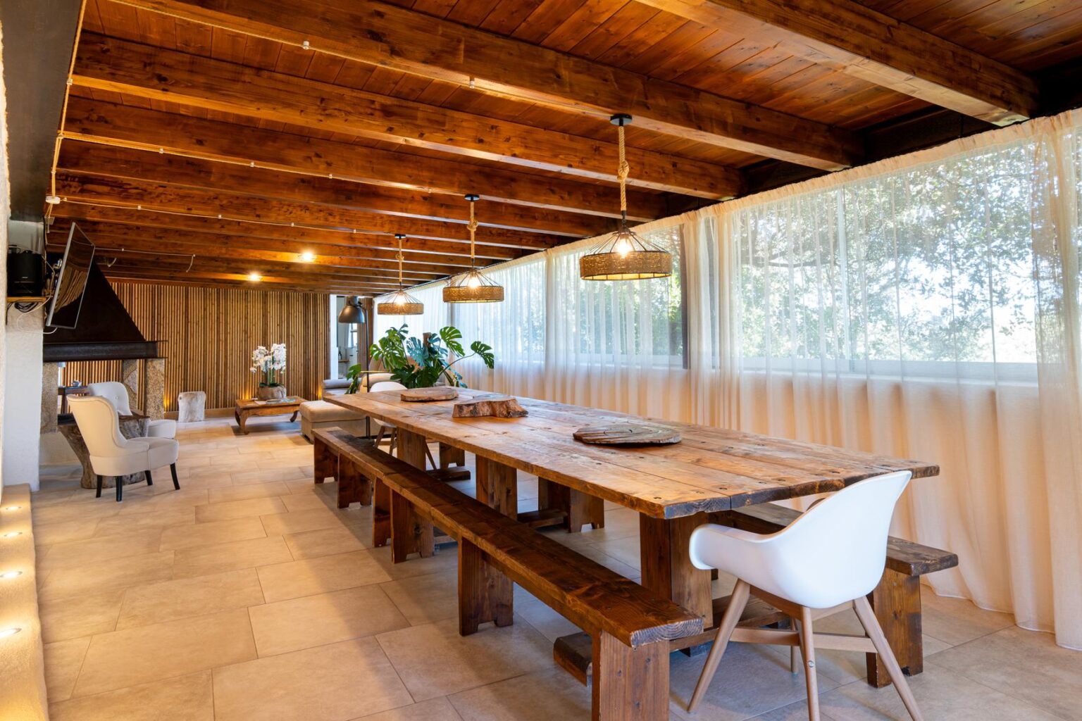 Spacious dining area featuring a large wooden table with bench seating, modern white chairs, and warm wooden beams overhead, illuminated by stylish pendant lights, creating a cozy and inviting atmosphere. | Only on SopranoVillas