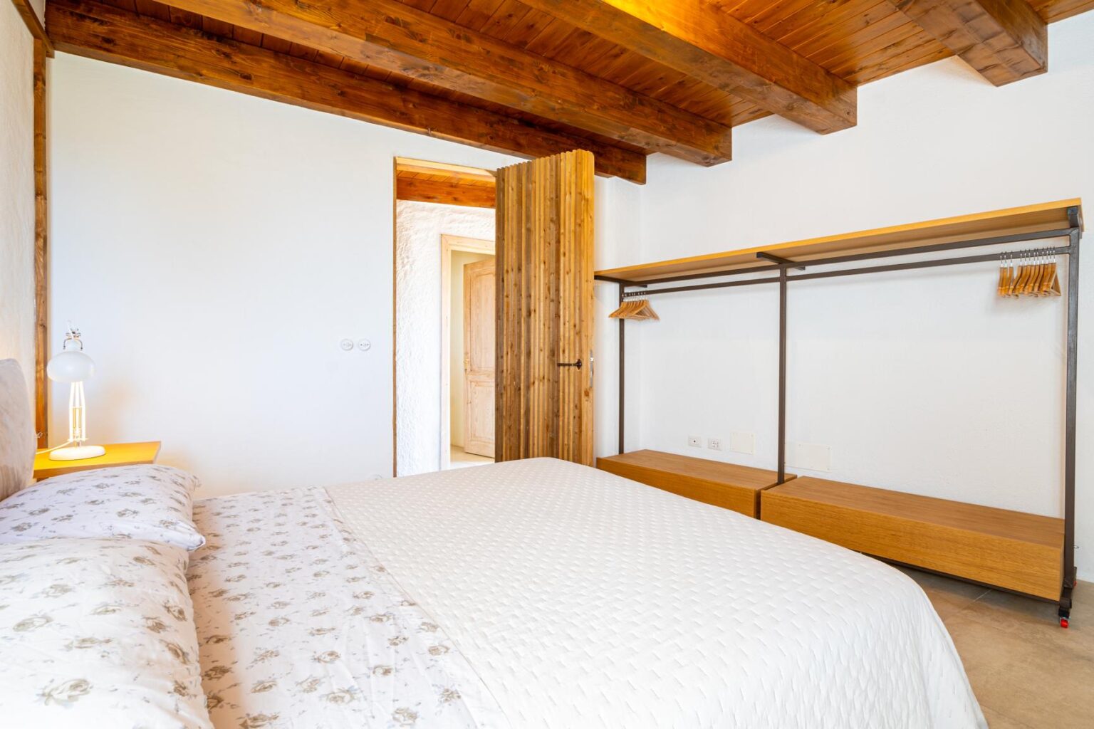 Modern bedroom interior featuring a double bed with a light-colored quilt, wooden beams on the ceiling, and minimalist wooden furniture including an open wardrobe and bedside lamp. Bright and airy atmosphere with neutral wall colors. | Only on SopranoVillas