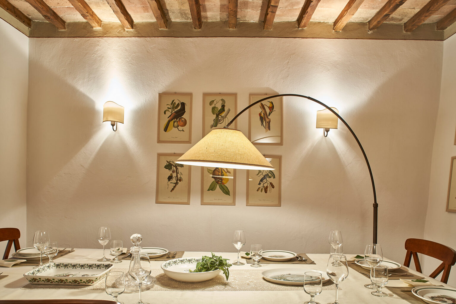 Elegant dining room featuring a beautifully set table with glassware and decorative plates, illuminated by a stylish arc lamp. The walls are adorned with vintage botanical art, creating a warm and inviting atmosphere perfect for entertaining guests. | Only on SopranoVillas
