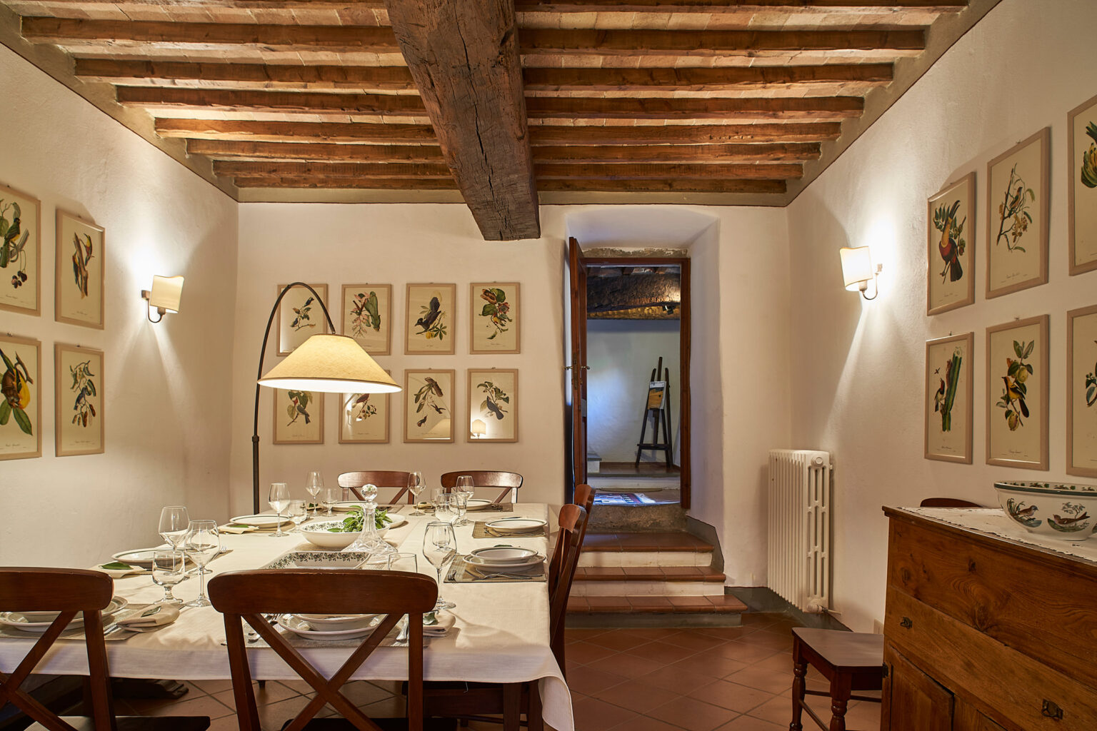 Cozy dining room featuring a wooden table set for a meal, adorned with elegant dinnerware and glassware. The walls are decorated with botanical prints, and warm lighting creates an inviting atmosphere. A doorway leads to another space, enhancing the room's charm. | Only on SopranoVillas