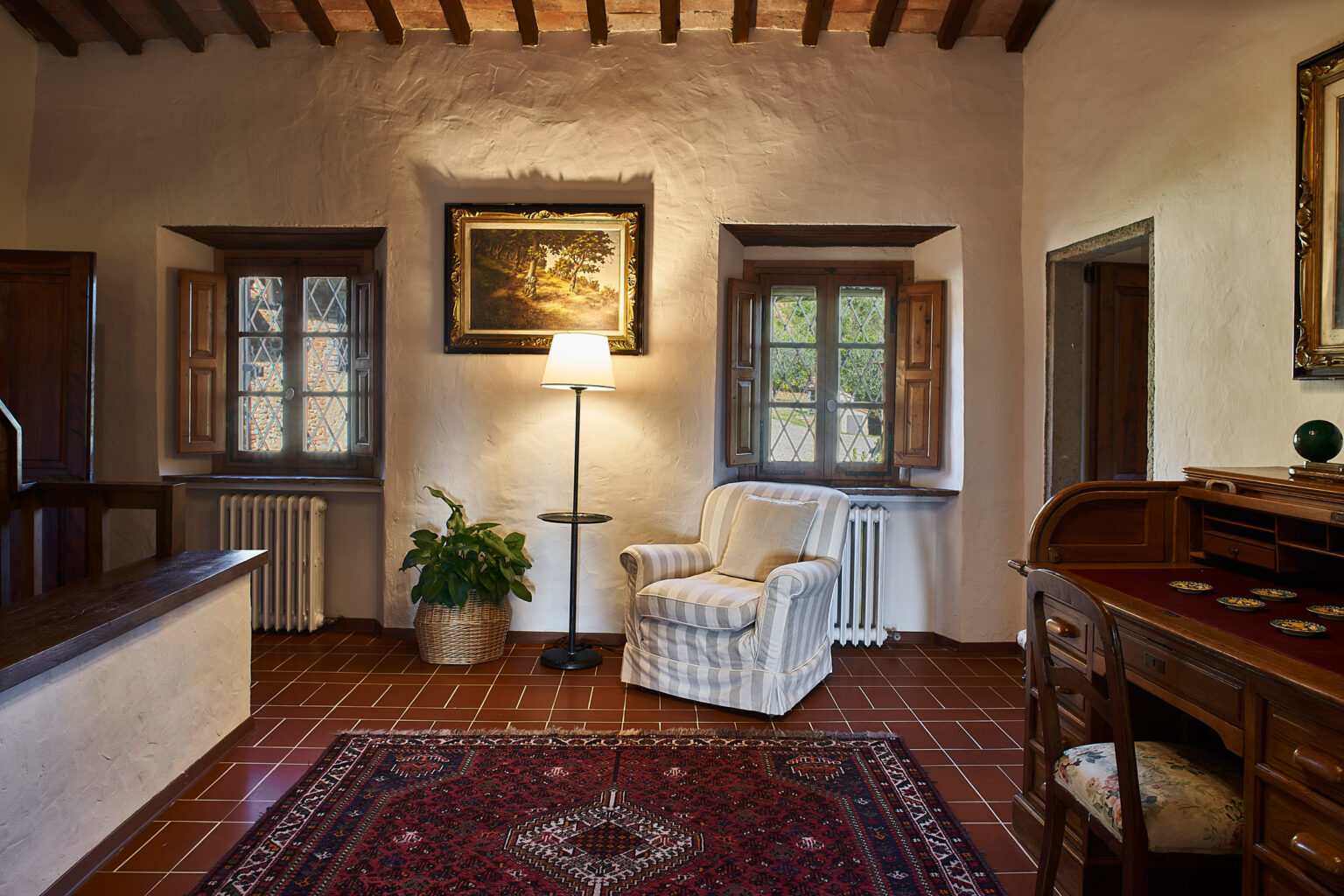 Cozy living room featuring a comfortable white armchair, a standing lamp, and wooden furniture. Natural light streams through window panes, highlighting the rustic decor and warm tones of the space. A decorative rug adds texture to the terracotta tiled floor. | Only on SopranoVillas