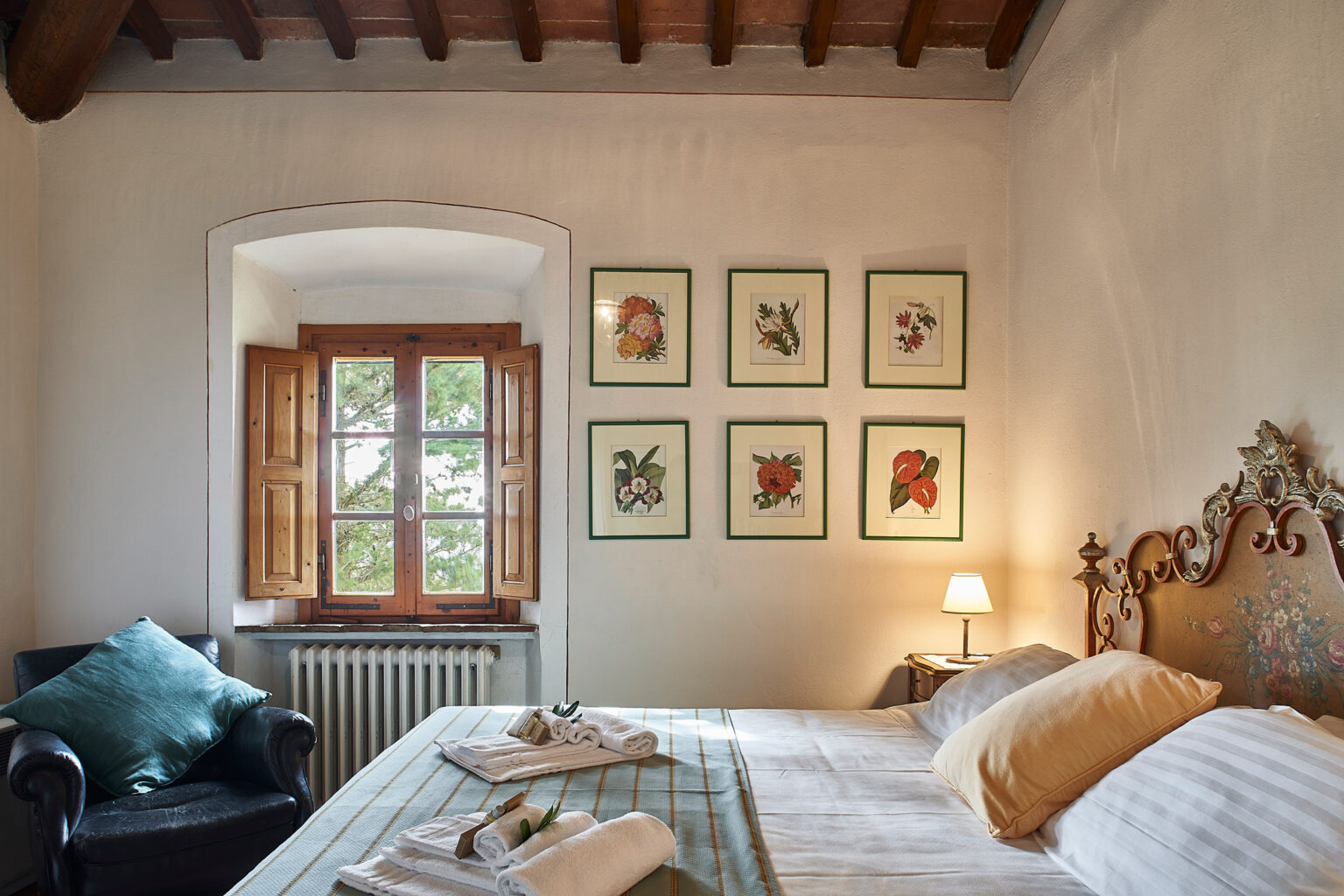 Cozy bedroom featuring a vintage wooden bed with decorative pillows, a comfortable armchair, and framed botanical prints on the wall. Natural light streams in through a window with wooden shutters, creating a warm and inviting atmosphere. | Only on SopranoVillas