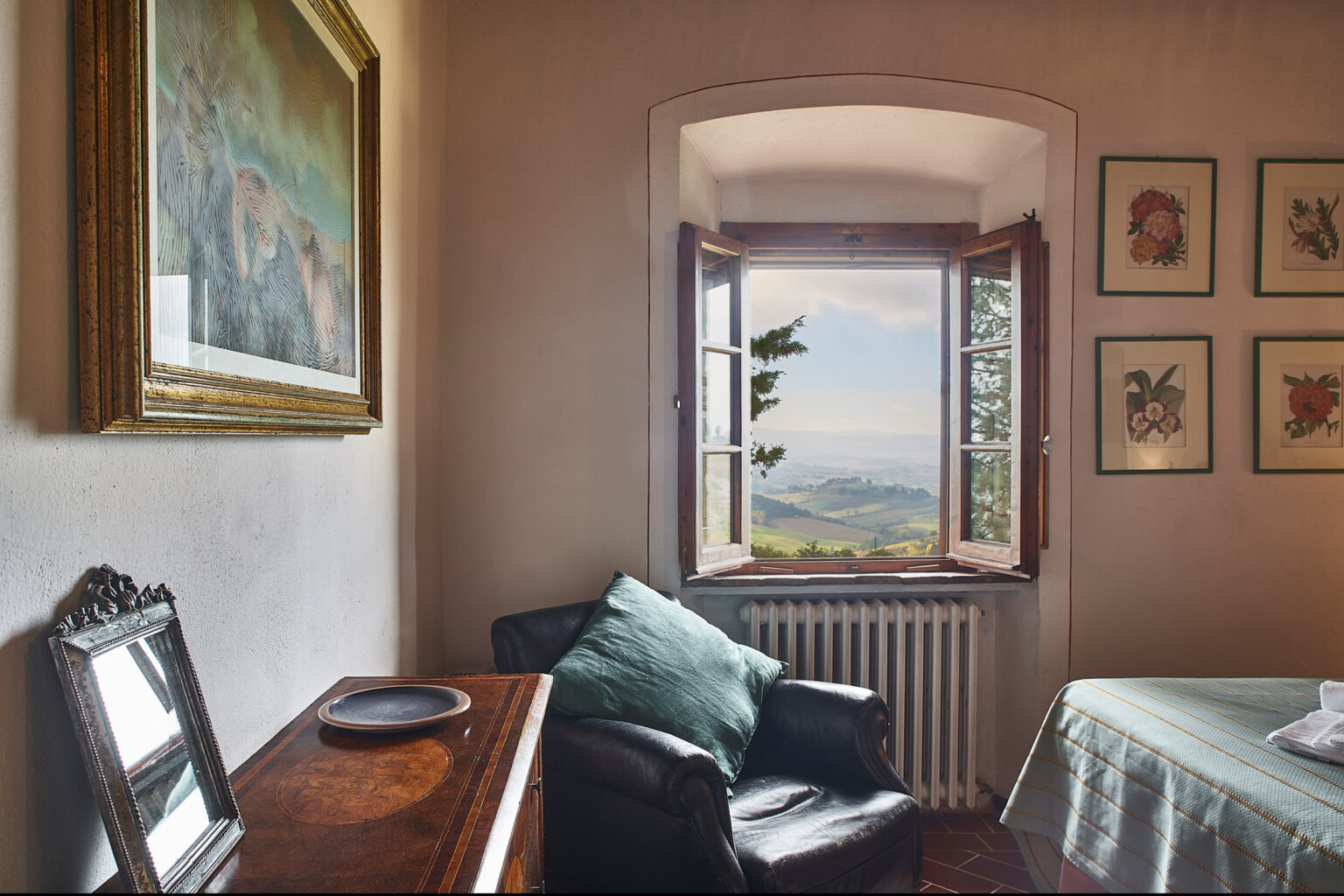 Cozy interior of a room featuring a comfortable chair, a wooden table, and decorative artwork on the walls, with a scenic view of rolling hills visible through an open window. | Only on SopranoVillas