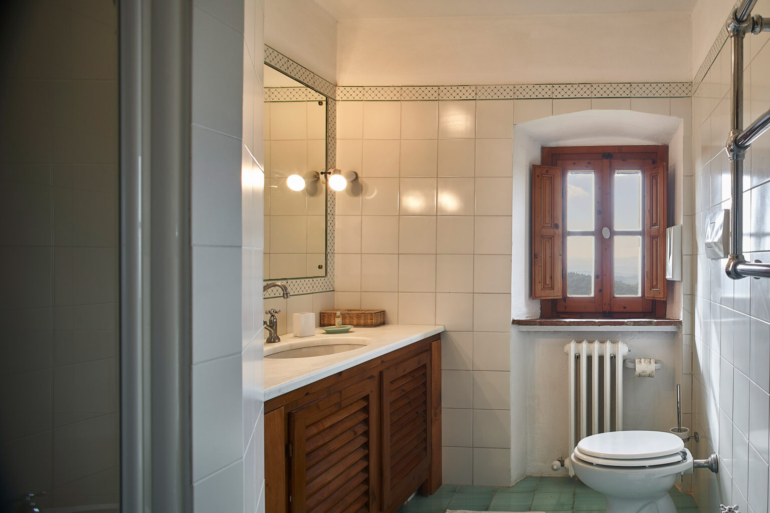 Modern bathroom featuring a wooden vanity, mirror, and window with traditional shutters, complemented by a toilet and radiator. The tile walls and natural light create a bright, inviting atmosphere. | Only on SopranoVillas