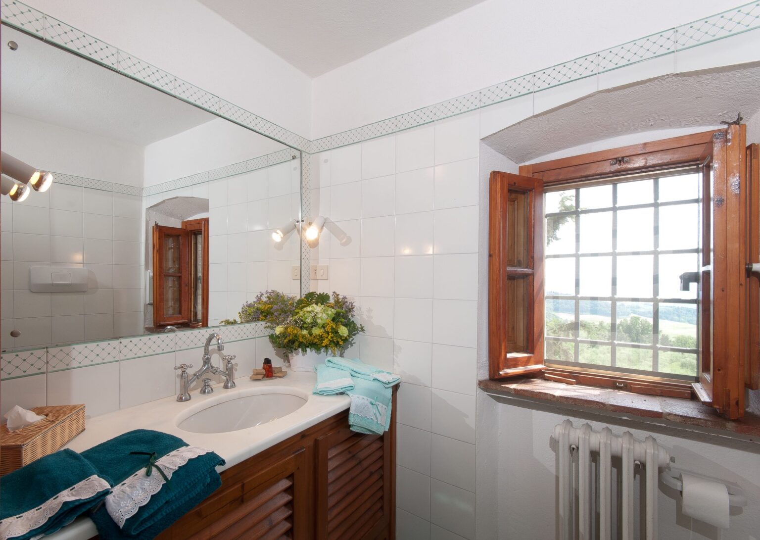 Bright and airy bathroom featuring a large mirror, wooden window frames, and a scenic view. The space includes a white sink, wooden cabinetry, and teal towels, creating a fresh and inviting ambiance. | Only on SopranoVillas