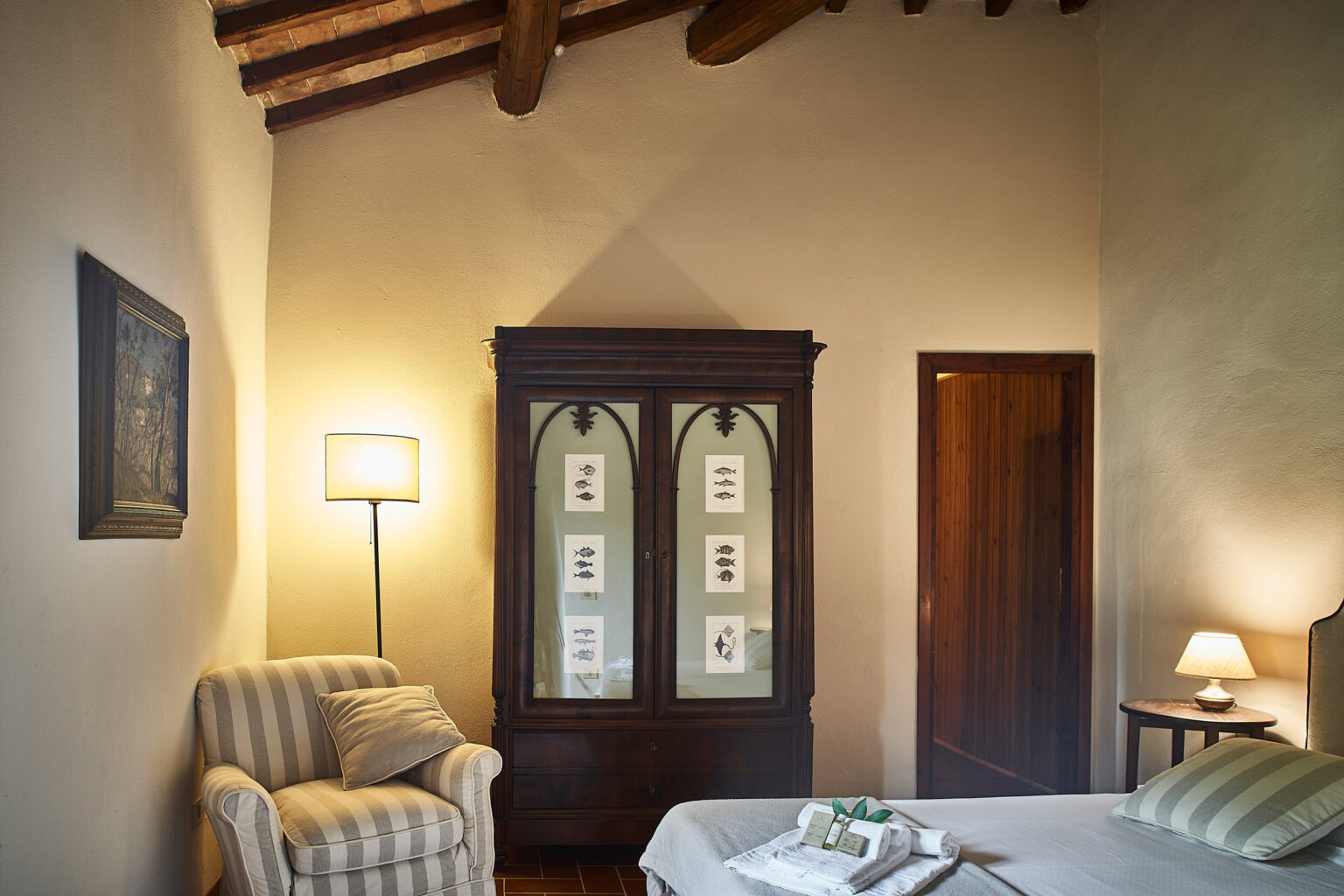 Cozy bedroom interior featuring a striped armchair, a wooden cabinet with framed art, and warm lighting from lamps, highlighting rustic decor and a serene atmosphere. | Only on SopranoVillas