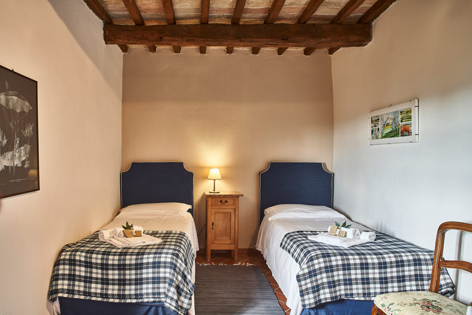 Cozy guest room featuring two single beds with blue headboards, a wooden nightstand with a lamp, and rustic wooden beams on the ceiling. The room is decorated with a plaid throw and wall art, creating a warm and inviting atmosphere. Ideal for travelers seeking comfortable accommodations. | Only on SopranoVillas