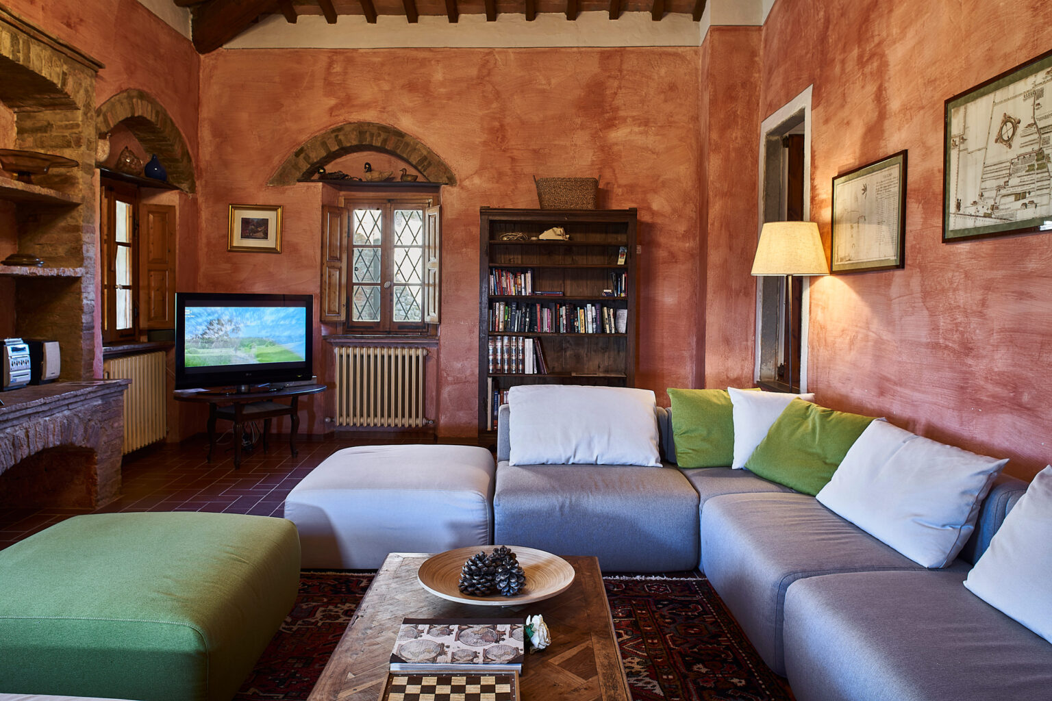 Cozy living room featuring a comfortable sectional sofa with green and white cushions, a wooden coffee table with decorative items, a TV displaying scenic imagery, and a bookshelf filled with books. The room has rustic charm with terracotta walls, wooden beams, and large windows allowing natural light. | Only on SopranoVillas