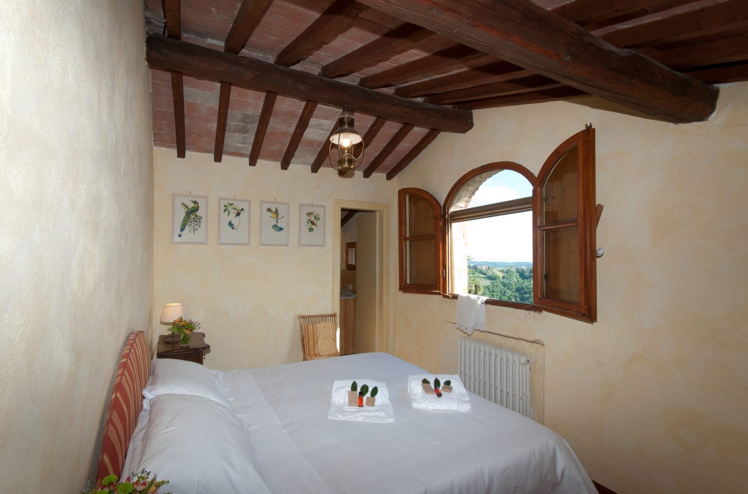 Cozy bedroom with a double bed, rustic wooden beams, and large windows offering scenic views. The room features soft bedding, decorative towels, and framed botanical prints on the walls, creating a warm and inviting atmosphere. Ideal for a relaxing getaway. | Only on SopranoVillas