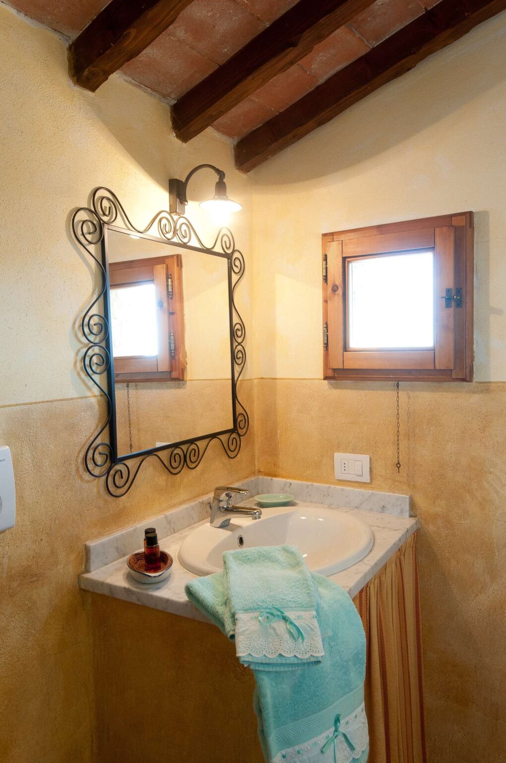Cozy bathroom interior featuring a marble sink, ornate black mirror, wooden window, and warm-toned walls. A turquoise towel hangs from the sink, adding a pop of color, while a small decorative bowl and toiletries complete the inviting space. | Only on SopranoVillas