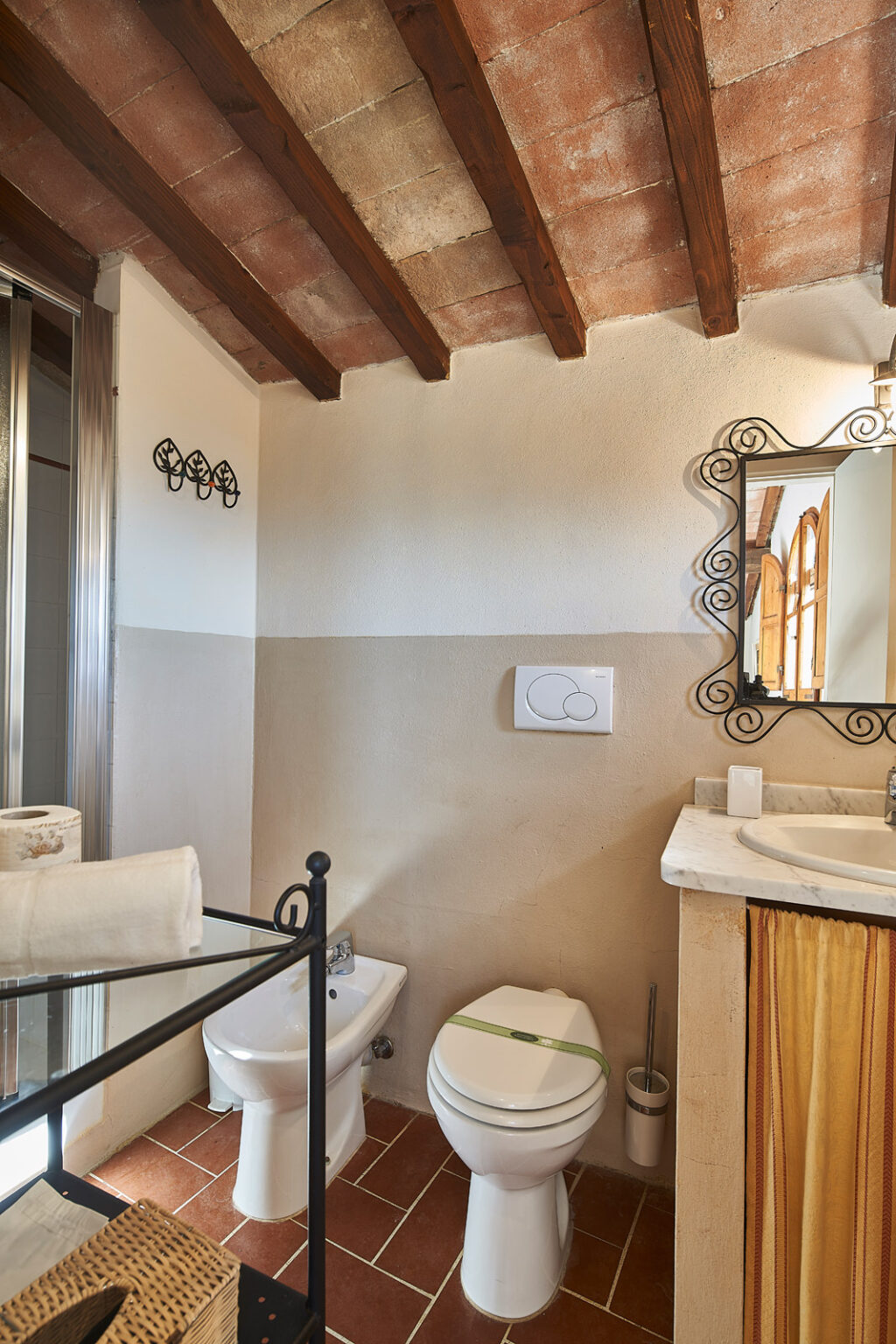 Cozy bathroom interior featuring rustic wooden beams, a bidet, and a modern toilet. The space includes a marble sink with a decorative mirror, towel rack, and subtle earthy tones on the walls. | Only on SopranoVillas