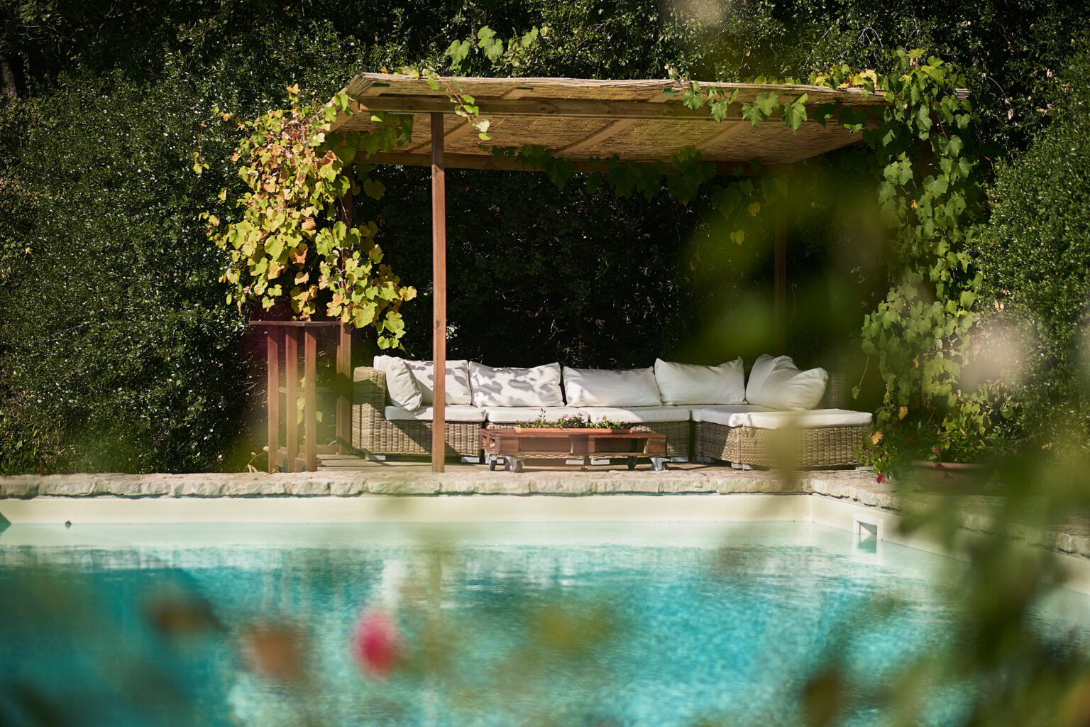 Cozy outdoor lounge area with a shaded pergola beside a swimming pool, surrounded by lush greenery and colorful flowers. Perfect for relaxation and summer gatherings. | Only on SopranoVillas