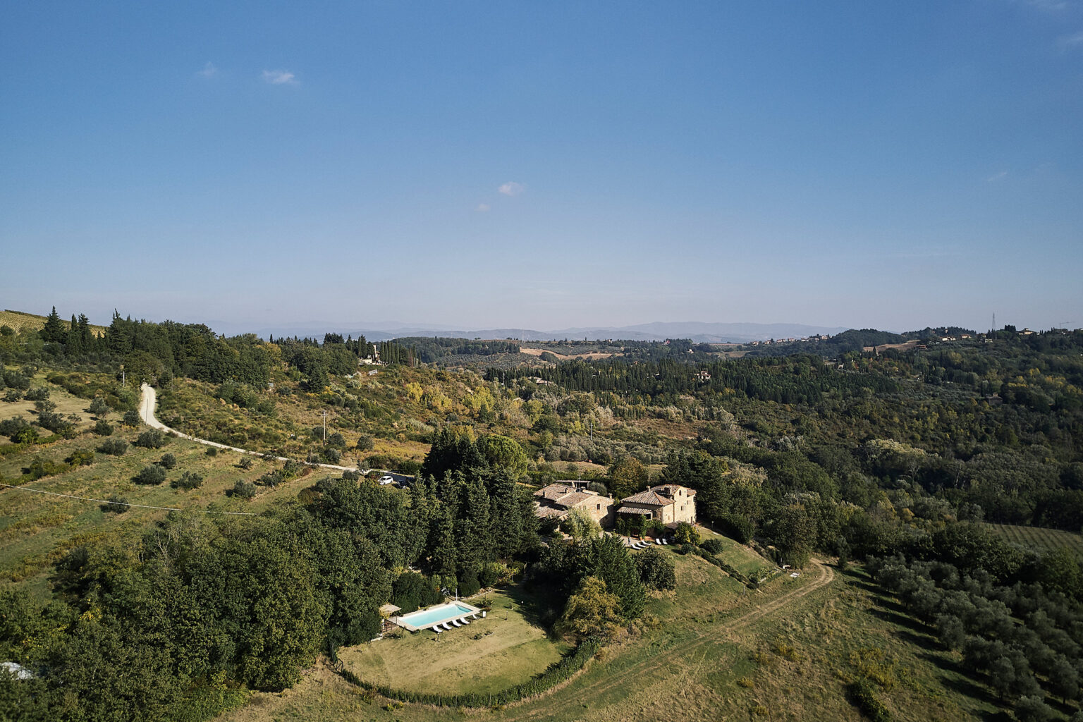 Aerial view of a rustic farmhouse surrounded by lush green hills and a swimming pool in the foreground, showcasing the scenic beauty of the Italian countryside under a clear blue sky. | Only on SopranoVillas