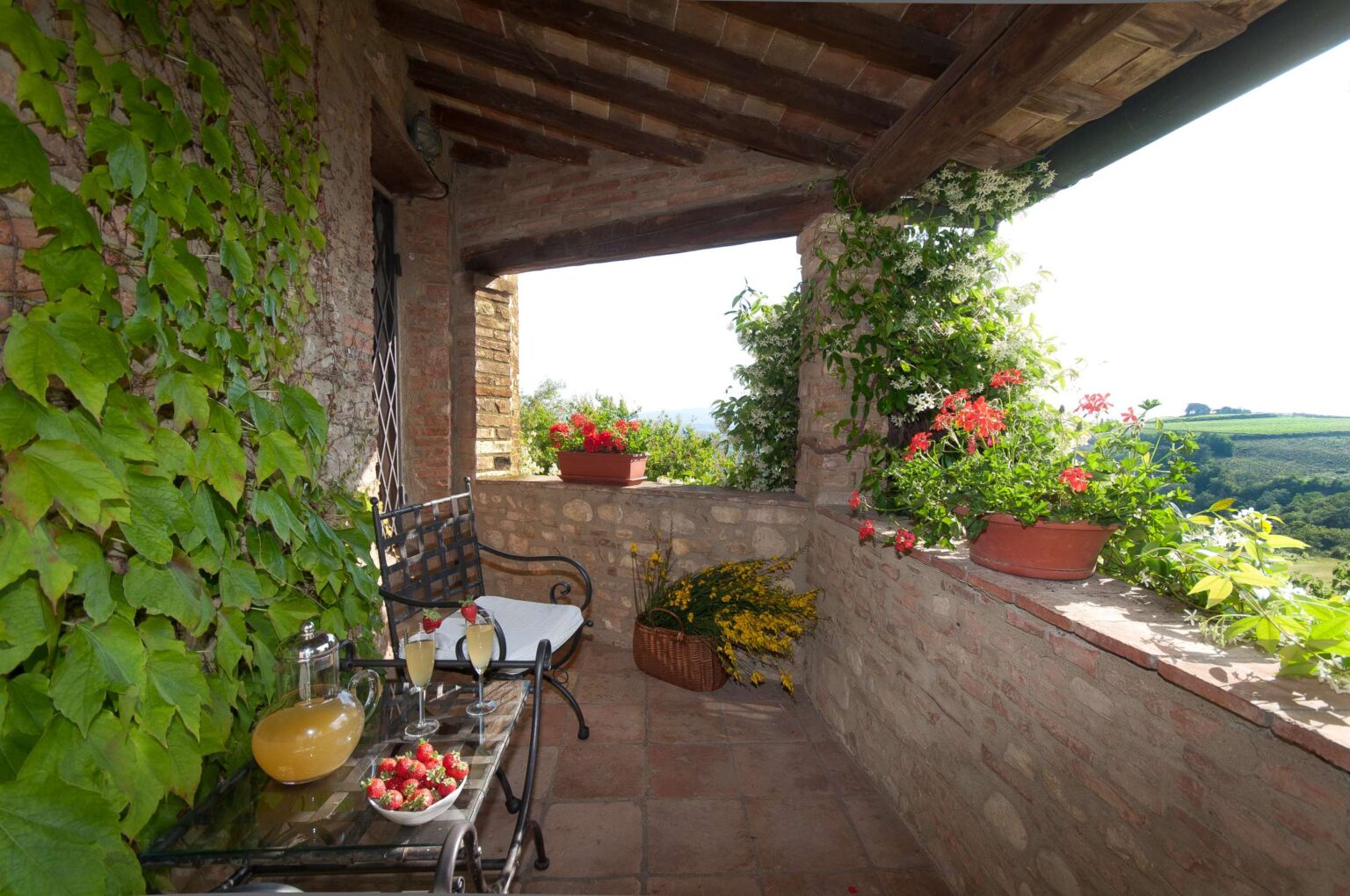 Charming terrace with a rustic stone wall adorned with vibrant green vines and colorful flowers, featuring a glass table set with a pitcher of lemonade, two glasses, and a bowl of strawberries, offering a scenic view of rolling hills in the background. | Only on SopranoVillas
