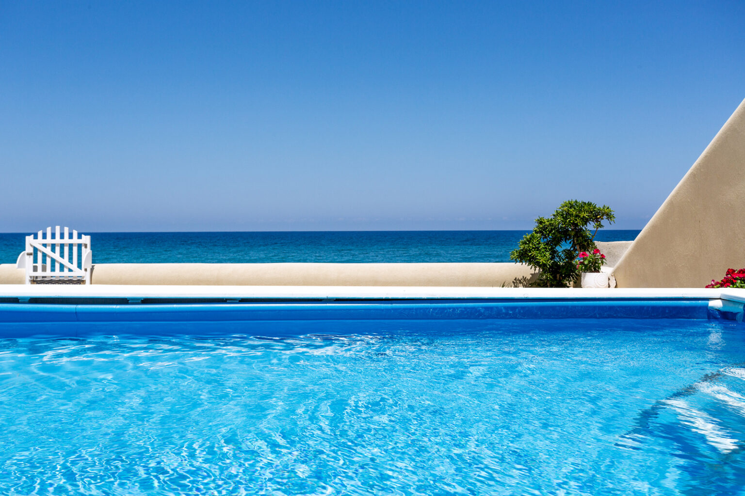 Clear blue swimming pool with a view of the ocean and a bright blue sky, featuring a white fence and tropical plants nearby. Ideal for vacation and relaxation. | Only on SopranoVillas
