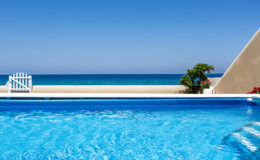 Clear blue swimming pool with a view of the ocean and a bright blue sky, featuring a white fence and tropical plants nearby. Ideal for vacation and relaxation.