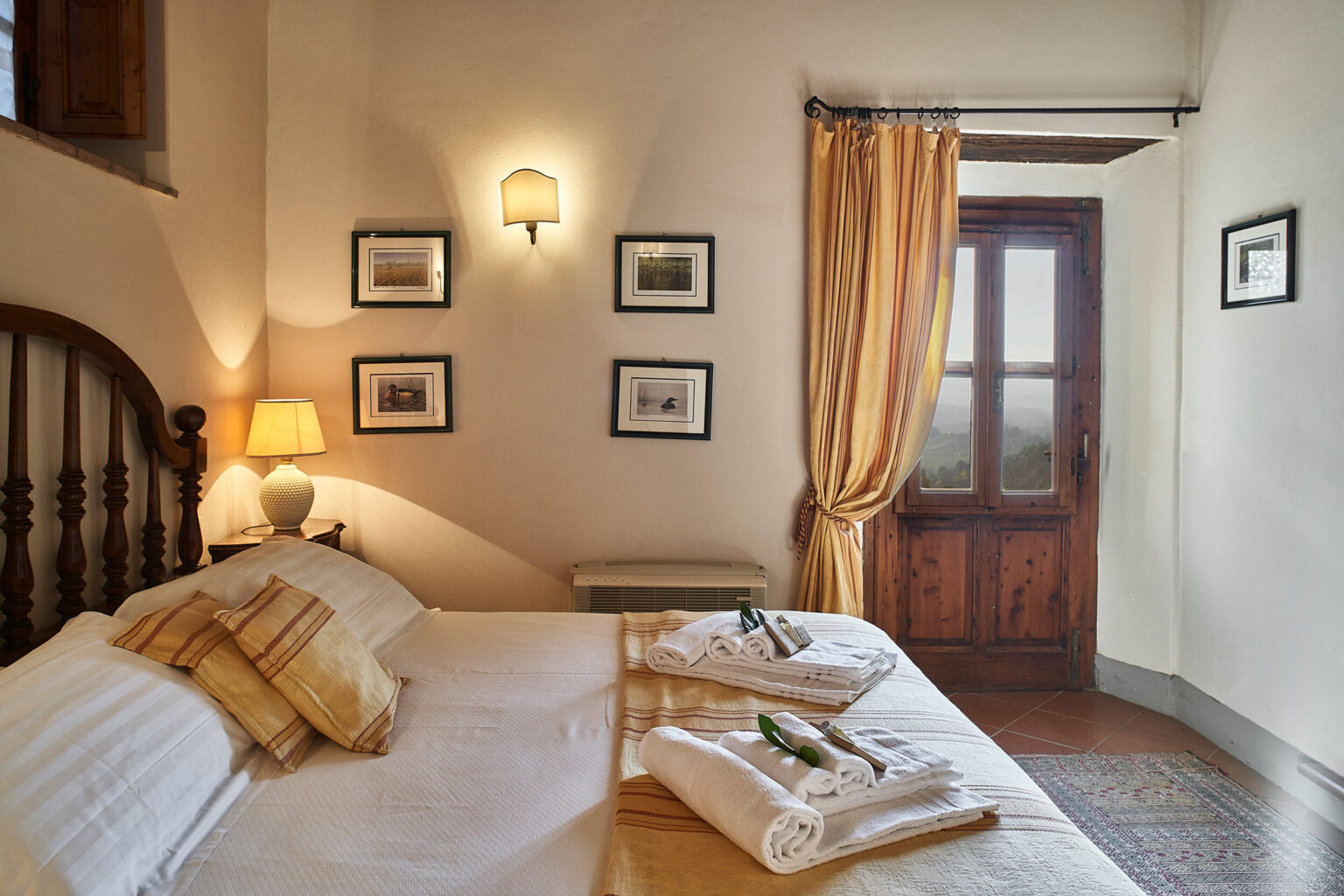 Cozy bedroom featuring a double bed with light-colored linens, decorative pillows, and neatly arranged towels. The room is illuminated by a bedside lamp and adorned with framed pictures on the walls. A window with yellow curtains offers a view of the outdoors, adding warmth to the inviting atmosphere. | Only on SopranoVillas