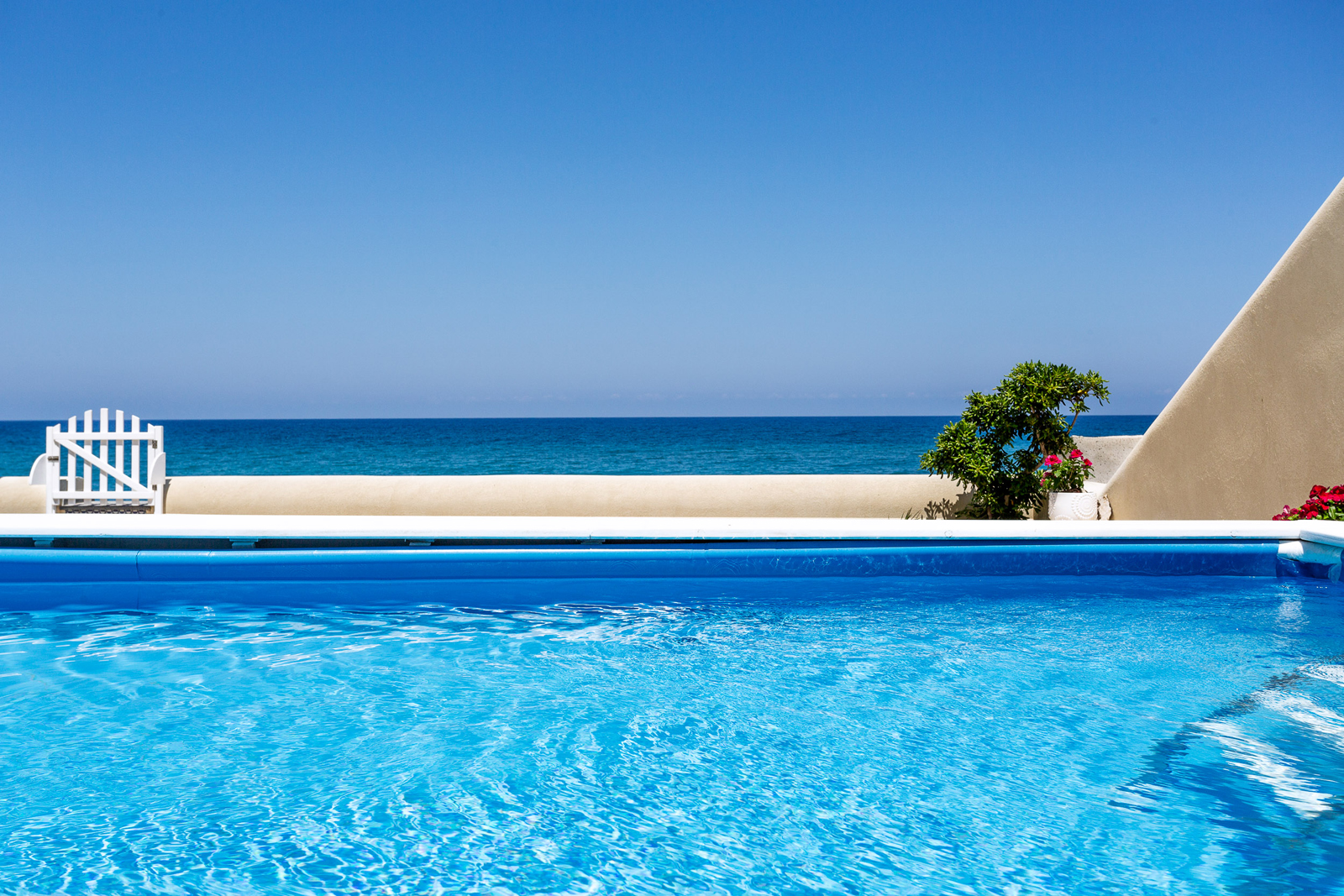 Clear blue swimming pool with a view of the ocean and a bright blue sky, featuring a white fence and tropical plants nearby. Ideal for vacation and relaxation. | Only on SopranoVillas
