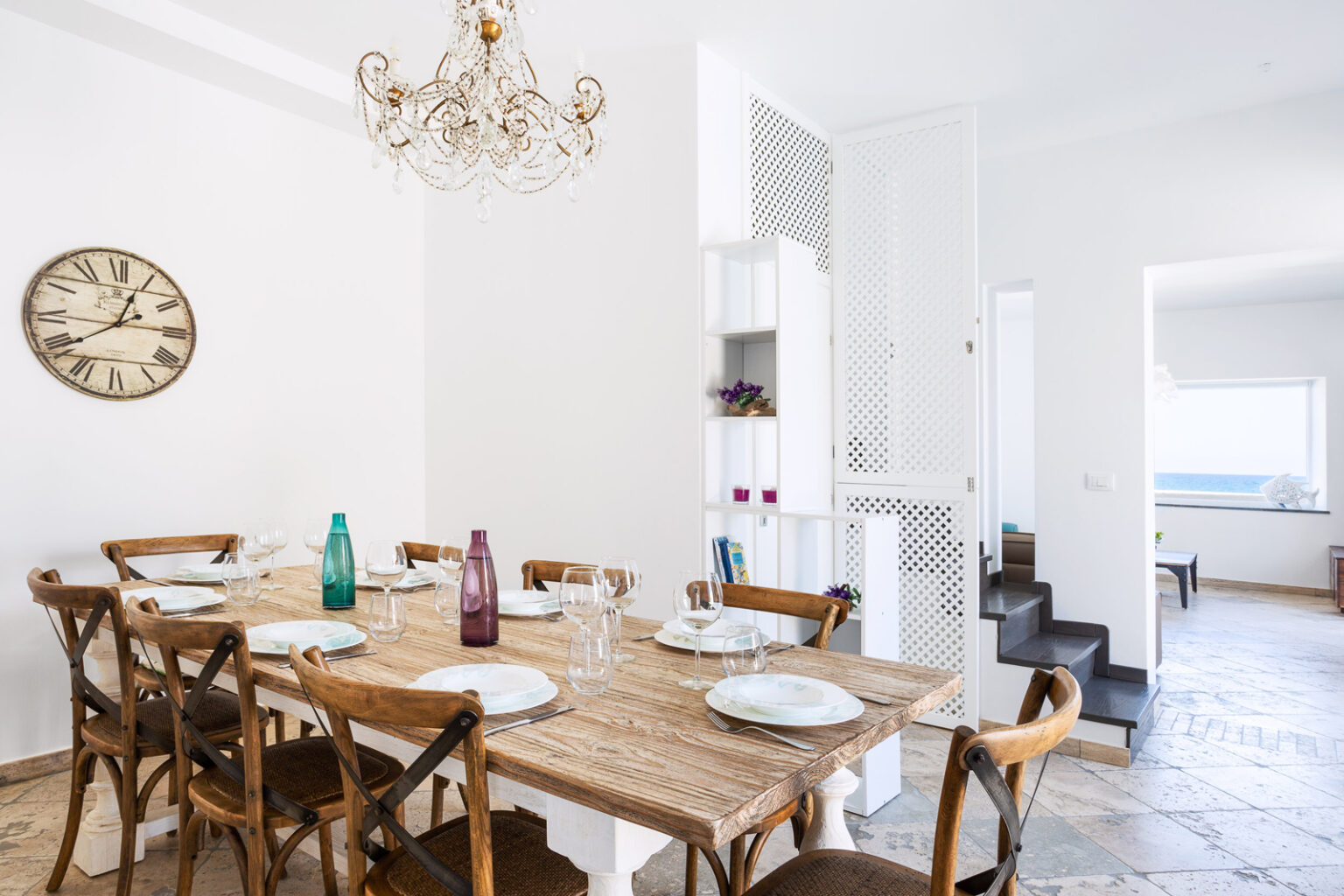 Bright and airy dining room featuring a rustic wooden table set for six, with elegant tableware and colorful glassware. A stylish chandelier hangs above, complementing the modern decor, while a clock adds a timeless touch. The open layout leads to a well-lit kitchen and living area, enhancing the inviting atmosphere. | Only on SopranoVillas