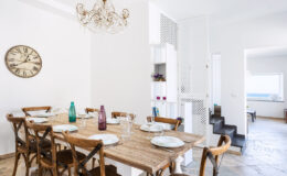 Bright and airy dining room featuring a rustic wooden table set for six, with elegant tableware and colorful glassware. A stylish chandelier hangs above, complementing the modern decor, while a clock adds a timeless touch. The open layout leads to a well-lit kitchen and living area, enhancing the inviting atmosphere.