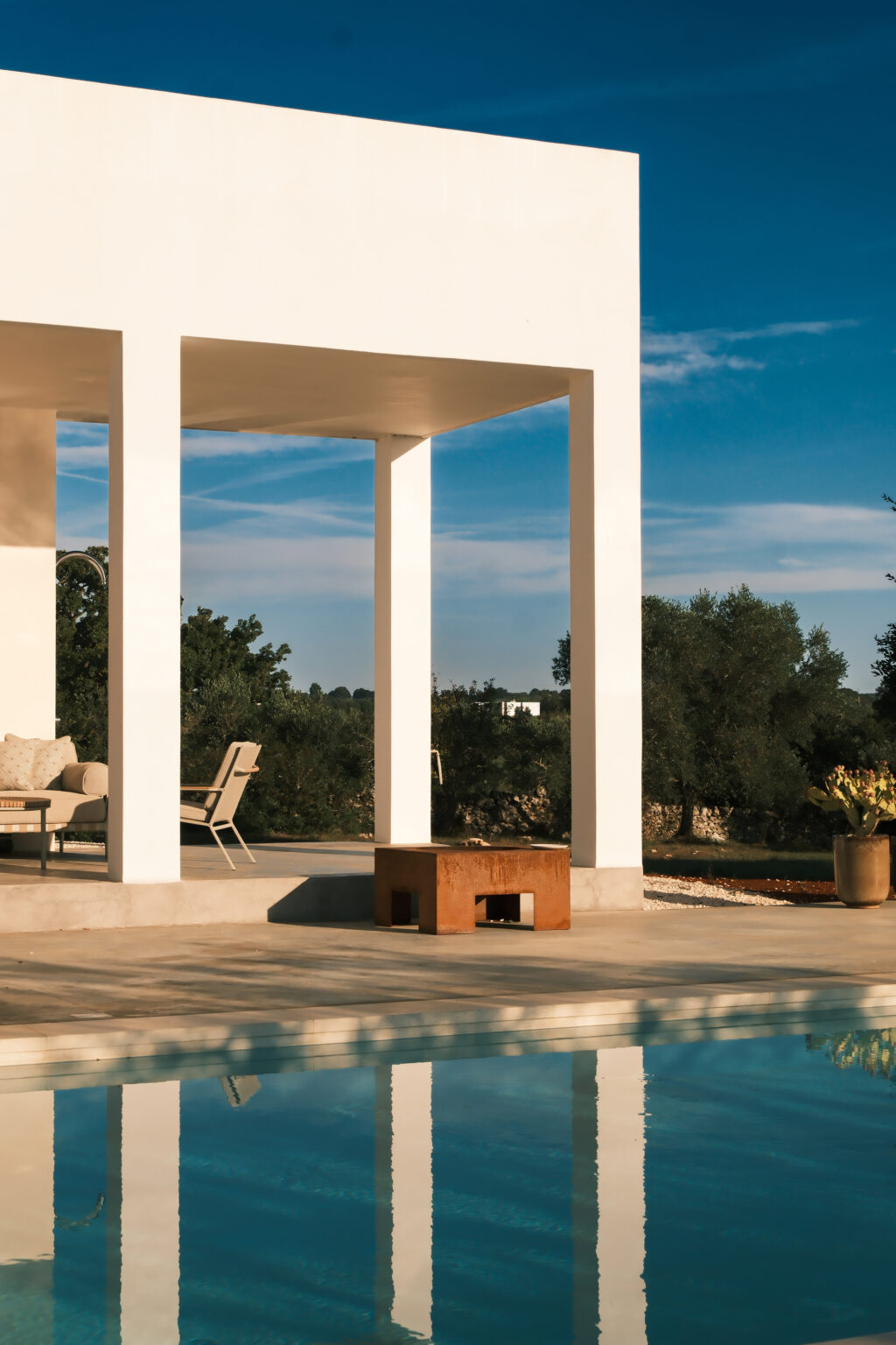 Modern outdoor living space featuring a minimalist design with white pillars, a rustic wooden table, and comfortable seating. Lush greenery and a clear blue sky create a tranquil atmosphere. | Only on SopranoVillas